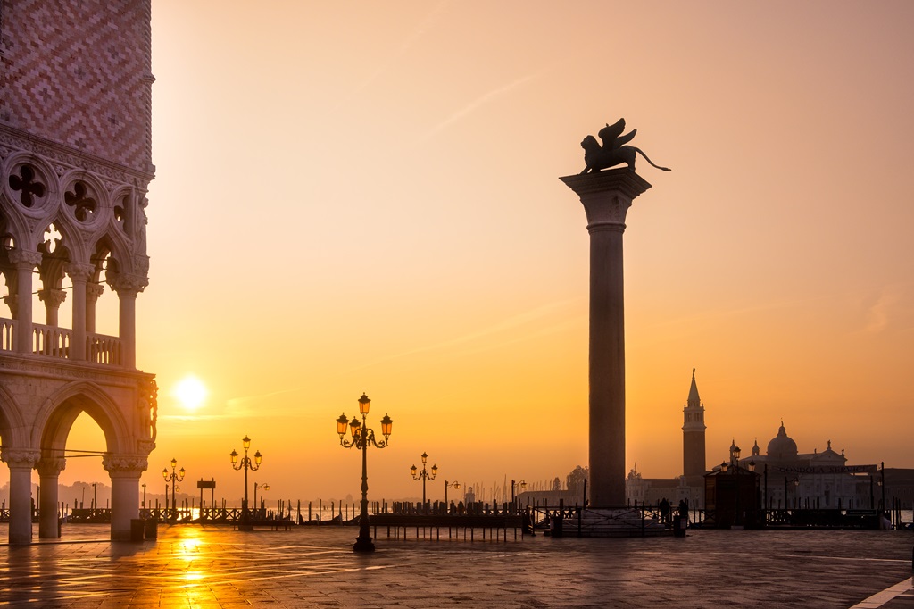 Alba dorata su Piazza San Marco a Venezia: vista con la Colonna del Leone di San Marco, Palazzo Ducale e il Campanile sullo sfondo, un'ispirazione per cosa vedere.