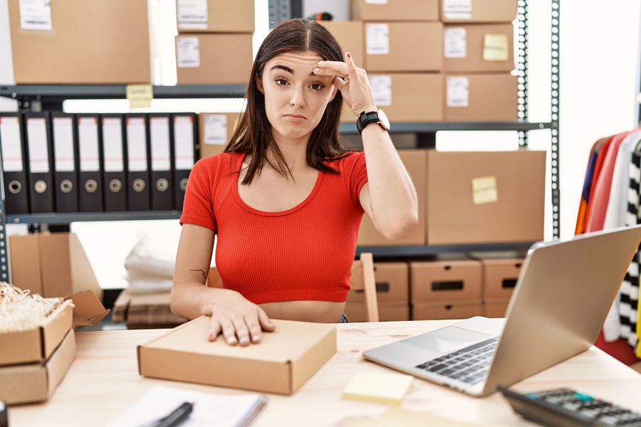 Giovane imprenditrice con espressione preoccupata al suo desk mentre si chiede come definire il prezzo di un prodotto, con pacchi e un laptop di fronte a lei.